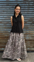 A woman modeling a sleeveless black button-up top paired with a long black and beige patterned skirt, standing in front of a weathered blue metal shutter.