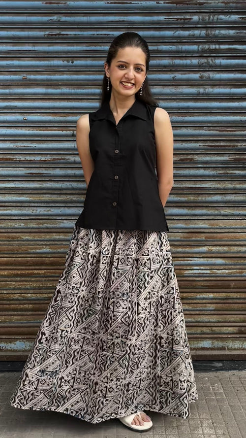 A woman modeling a sleeveless black button-up top paired with a long black and beige patterned skirt, standing in front of a weathered blue metal shutter.