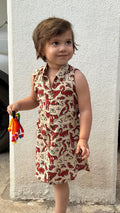 A toddler girl wearing a sleeveless collared dress with a red flamingo print on a beige background, standing outdoors next to a white pillar.
