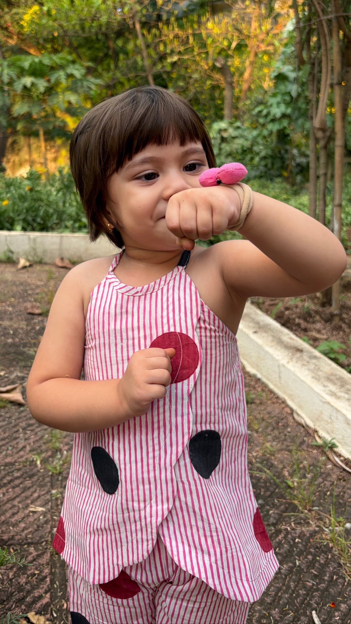 A toddler girl wearing a pink and white striped sleeveless top with black polka dots stands outdoors in a garden, holding a pink toy near her face.